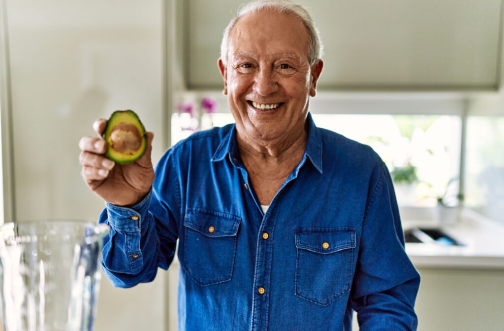 A senior man  is holding a n avocado cut in half and preparing an avocado shake in the kitchen.