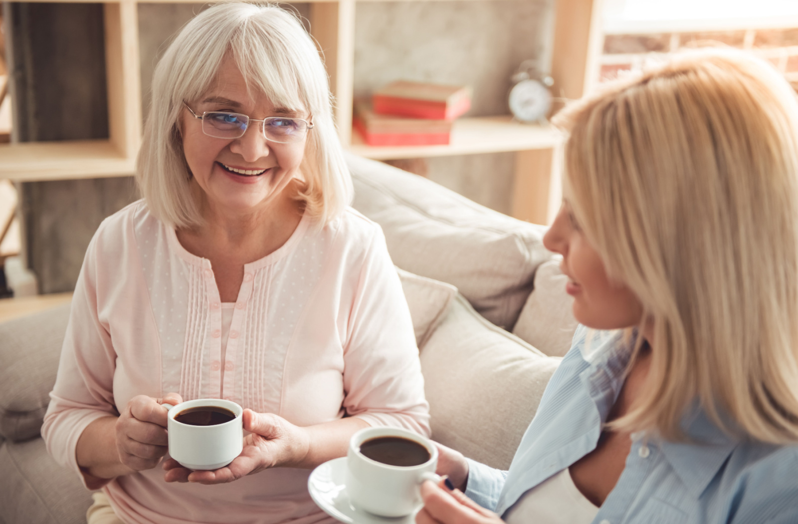 An older adult woman and her daughter sitting on a couch smiling and talking to each other while holding mugs of coffee