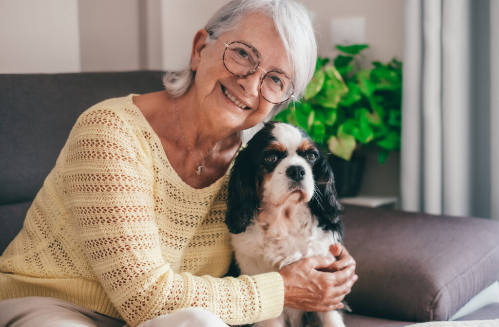 An older adult woman sitting on a couch with a dog, looking directly at the camera and smiling