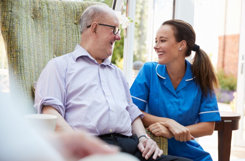 An older adult man in a memory care facility sitting on a chair smiling and having a conversation with a nurse.